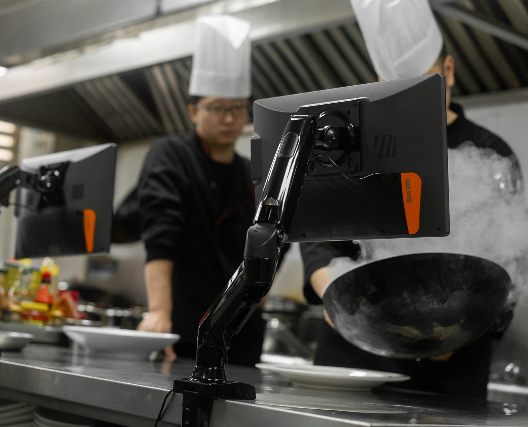 A busy commercial kitchen with two display screens on arms above the pass, chefs working a wok
