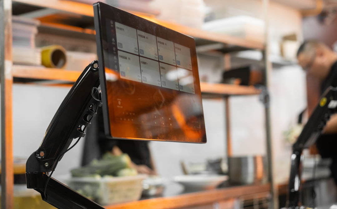 The kitchen display on an arm showing a grid of order tickets, kitchen shelving behind