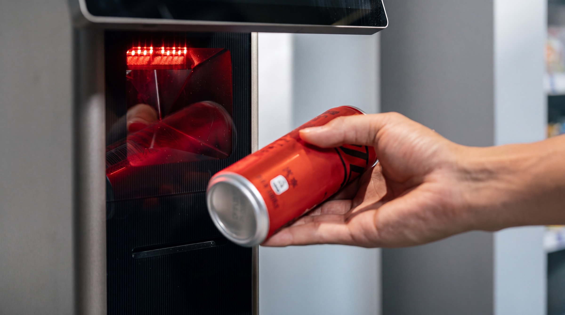 A hand holding a can to the barcode scanner set into a kiosk, the scanner's red lines on the glass