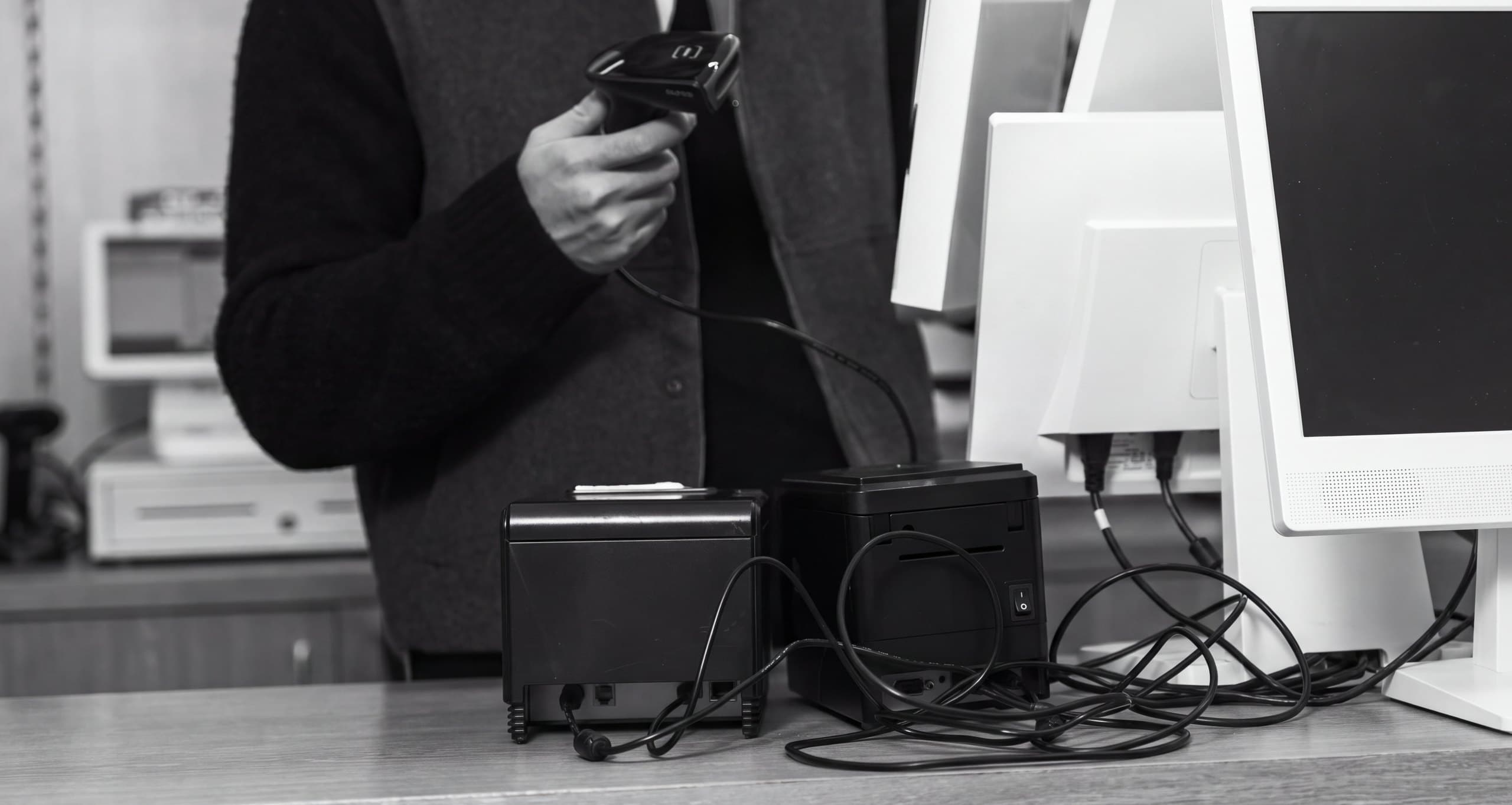 A shop counter in black and white: two countertop printers side by side among monitors, a hand-held scanner and their cabling